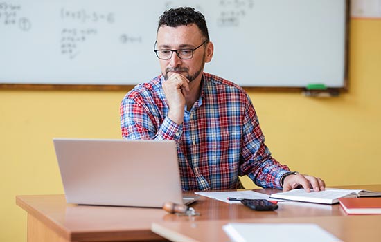 一位男老师在白板前的桌子上用笔记本电脑看书